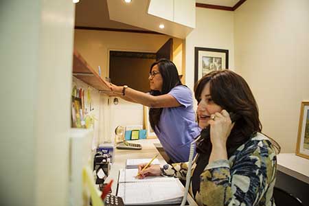 The image depicts two individuals, likely professionals, working at an office desk with papers and a computer monitor visible.