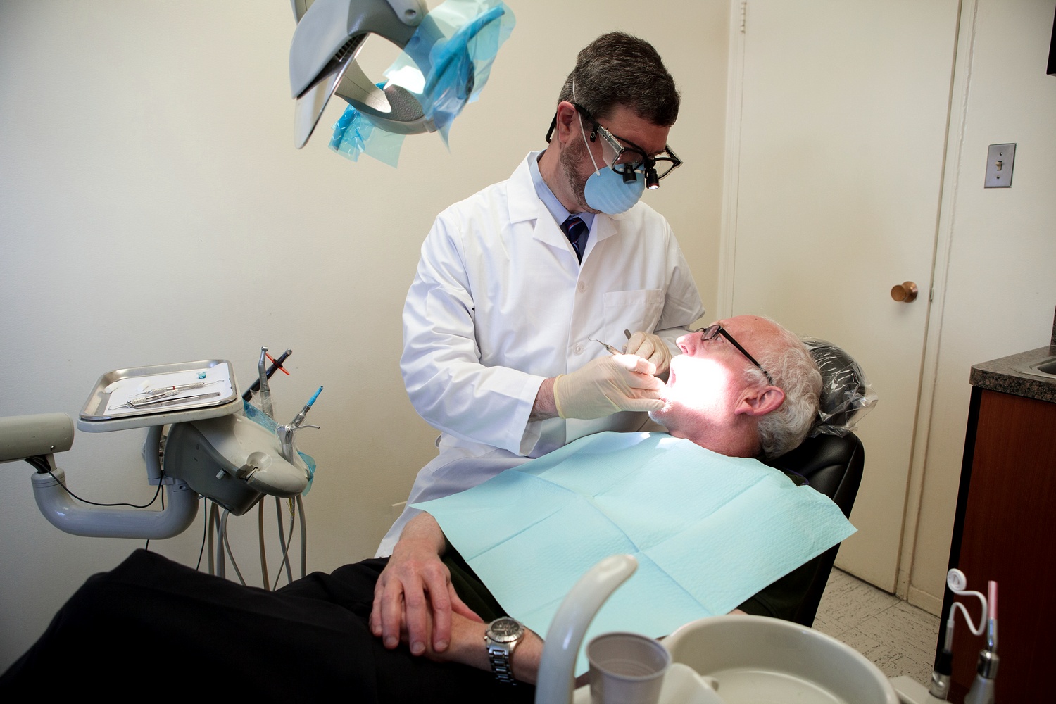 A dentist wearing glasses is performing dental work on an older man who is sitting in a chair with his eyes closed.