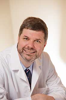 The image shows a man wearing a white lab coat, smiling at the camera, with a professional setting behind him, likely a workplace or office environment.