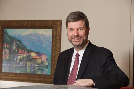 The image shows a man standing next to a framed picture, wearing a suit with a tie, smiling slightly, and looking directly at the camera.
