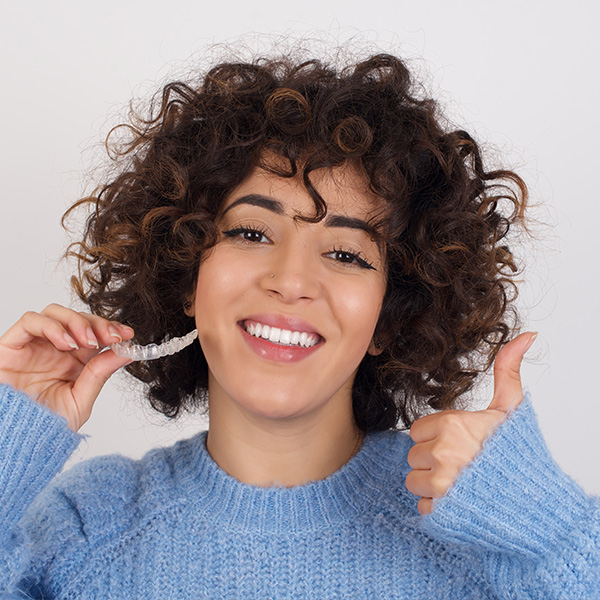 Woman with curly hair, blue sweater, and thumbs-up gesture, smiling at camera.
