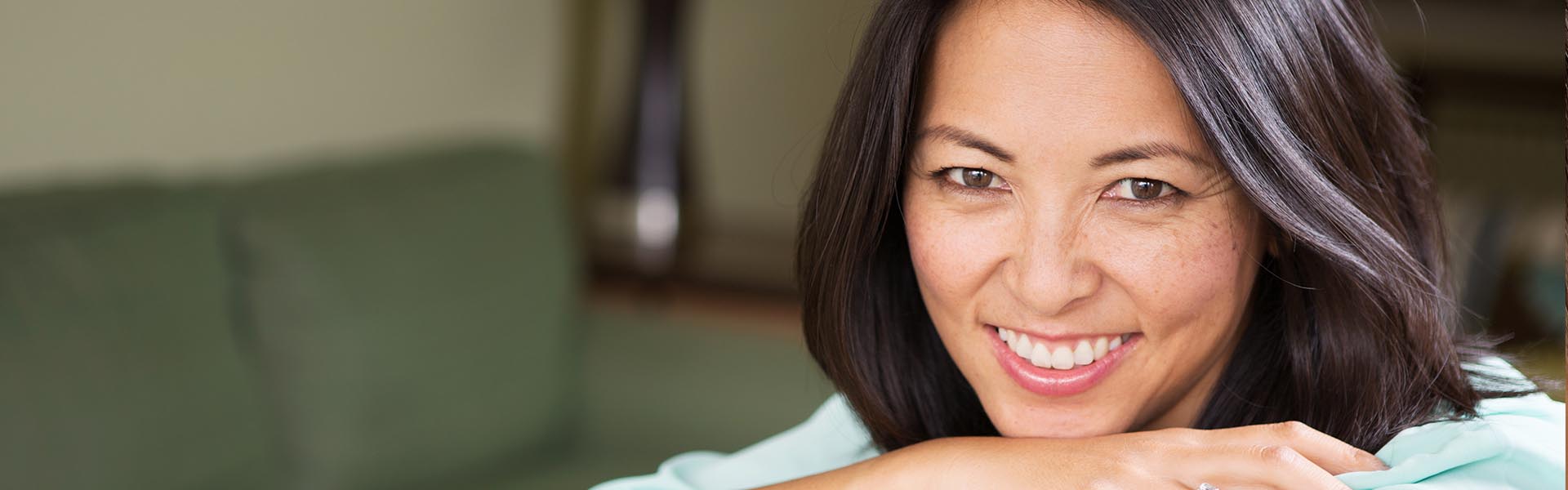 The image shows a woman with her hand on her chin, smiling at the camera, set against an indoor background with a couch visible.