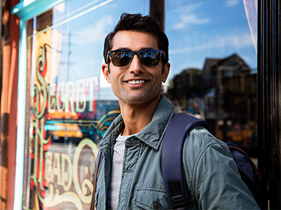 The image shows a man wearing sunglasses, standing outside a storefront with a sign that reads  Secret Life,  dressed in casual attire including a backpack.