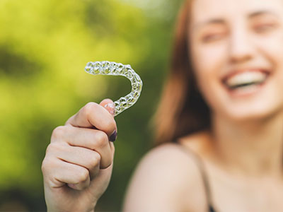 A smiling woman holds up a clear plastic toothbrush with bristles against a blurred outdoor background.