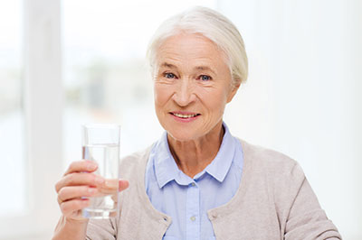 The image shows an elderly woman holding a glass of water, smiling, with a clear focus on her action.