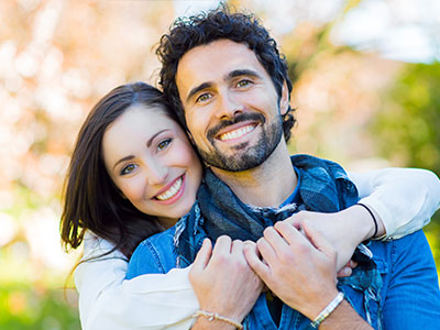 This is a color photograph depicting two adults, likely a man and a woman, embracing each other with smiles on their faces against a blurred outdoor background that suggests a sunny day.