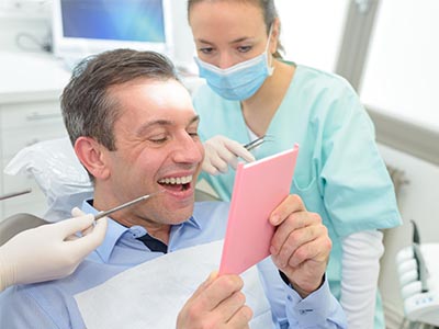 In the image, there is a man sitting in a dental chair, holding a pink card with a surprised expression, while a dental professional attends to him.