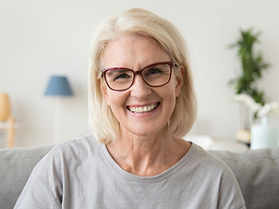 A smiling woman wearing glasses and a light-colored top, seated indoors with a white wall in the background.