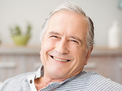 The image shows an elderly man with gray hair, smiling at the camera, wearing a blue shirt, seated comfortably in a home setting.