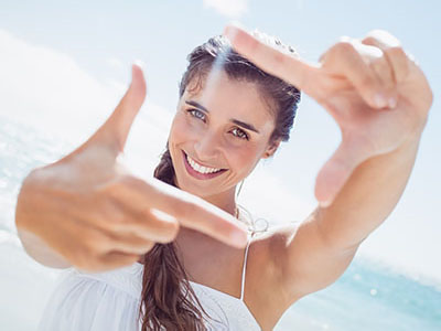 A smiling woman with long hair holds up her hand in front of her face, creating a frame with her fingers, against a sunny beach backdrop.