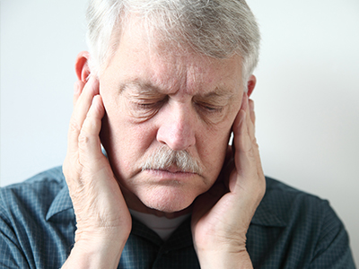 The image shows an elderly man with his eyes closed, holding his face with both hands near his ears, appearing to be asleep or resting with a pained expression.